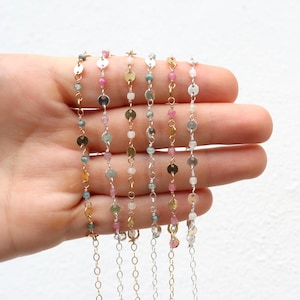 May include: A collection of delicate necklaces with colorful gemstones and small, round, metallic accents. The necklaces feature a variety of colors, including pink, green, and gold, and are displayed against a neutral background.
