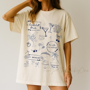 Op de afbeelding: Crèmekleurig T-shirt met blauwe illustraties en tekst. Het ontwerp bevat cocktailglazen, een discobal en de zinnen "a bombshell Bride" en "Olivia's Bachelorette". Het shirt heeft ook de tekst "the villa has entered" en "September 2025 Miami, FL".