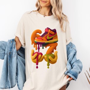 May include: Cream-colored t-shirt featuring a colorful graphic of a sneaker with the numbers "67" and dripping paint. The sneaker design incorporates orange, yellow, purple, and green hues. The graphic is centered on the shirt.
