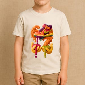 May include: A cream-colored t-shirt featuring a colorful, knitted graphic of a sneaker, the number 67, and hands with dripping paint. The sneaker has orange, yellow, purple, and green accents. The number 67 and hands are yellow, orange, and green.