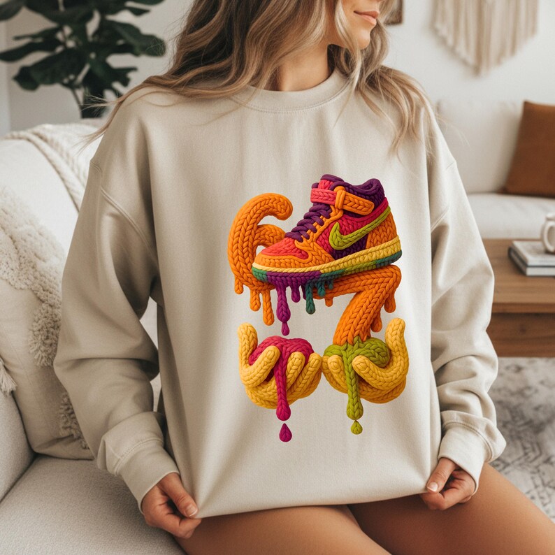May include: Beige sweatshirt featuring a colorful, artistic rendering of a high-top sneaker. The sneaker design incorporates orange, yellow, green, purple, and pink hues, with a dripping effect. The design is placed on the front of the sweatshirt.
