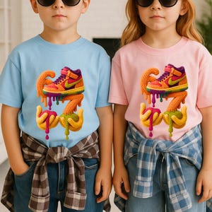 May include: Two children's t-shirts, one light blue and one pink, each featuring a colorful graphic of a sneaker with dripping accents. The sneakers are orange, purple, and yellow. The shirts are paired with blue jeans.