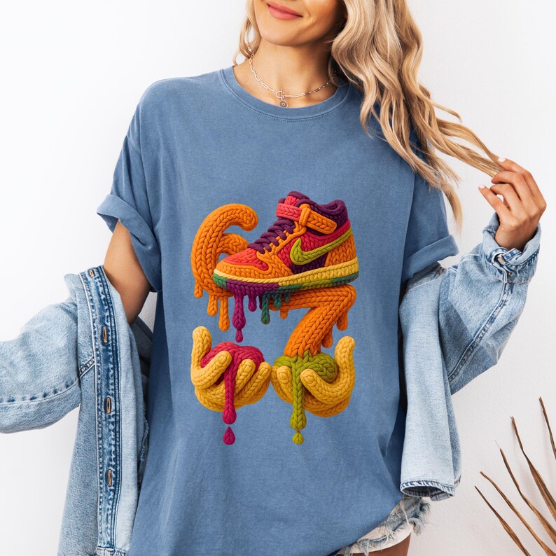 May include: A light blue t-shirt featuring a colorful, knitted sneaker design with dripping paint effect. The sneaker is in shades of orange, purple, yellow, and green. The design includes the number 67 and two hands.
