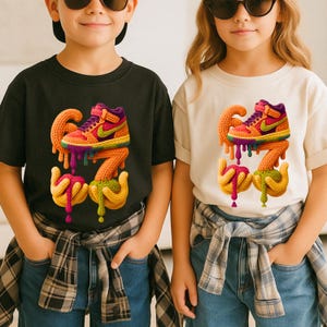 May include: Two children wearing black and cream-colored t-shirts. Each shirt features a colorful, cartoon-style sneaker design with the number "67" in a dripping, textured style. The sneakers have orange, purple, green, and yellow accents.
