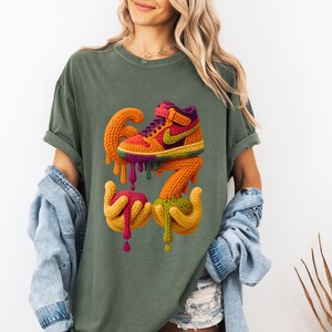 May include: Olive green t-shirt featuring a colorful, artistic rendering of a high-top sneaker with dripping paint. The design includes the number "67" and hands holding the dripping paint. The sneaker has a rainbow color scheme with a purple collar and a green Nike swoosh.