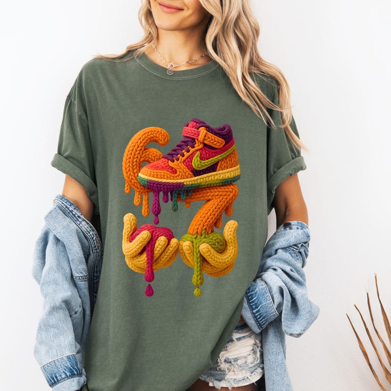 May include: Olive green t-shirt featuring a colorful, artistic rendering of a high-top sneaker with dripping paint. The design includes the number "67" and hands holding the dripping paint. The sneaker has a rainbow color scheme with a purple collar and a green Nike swoosh.