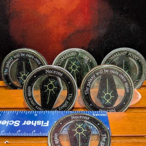 Peut inclure: Plusieurs jetons Necrons circulaires avec des détails scintillants. Chaque jeton présente un motif de cercueil noir avec un symbole vert, le mot "Necrons" et le texte "The galaxy will be ours once again". Une règle bleue est au premier plan.