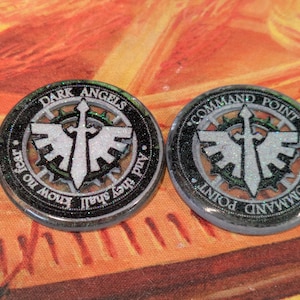 May include: Two round tokens with a black border and silver glitter accents. One token reads "DARK ANGELS" and "know no fear and they shall". The other reads "COMMAND POINT". Both feature a sword and wing design.