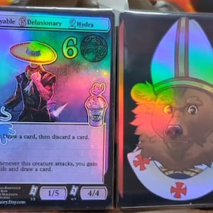 Puede incluir: Dos cartas coleccionables. La primera carta presenta una figura con un sombrero y un vaso de refresco, con el texto "Playable", "Delusionary" e "Hydra". La segunda carta muestra un oso con una mitra. Ambas cartas tienen efectos holográficos.