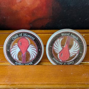 May include: Two round tokens with a brown and black border. Each token features a red drop shape with white wings and the text "Oath of Moment" and "Blood Angels". The tokens are on a wooden surface.