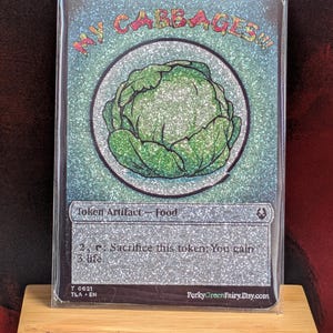 May include: A collectible card with a glittery background and a green cabbage illustration. The card reads "My Cabbages!!" at the top and includes text about the card's function as a token artifact. The card is in a protective sleeve.