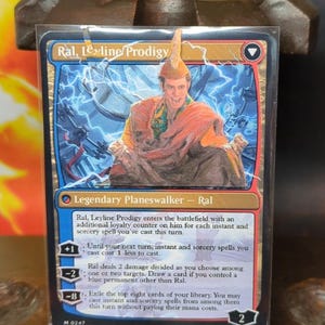 Puede incluir: Carta de Magic: The Gathering con Ral, Leyline Prodigy. Borde azul y dorado, ilustración de un hombre con túnicas naranjas. El texto incluye el nombre de la carta, sus habilidades y el símbolo de Planeswalker.