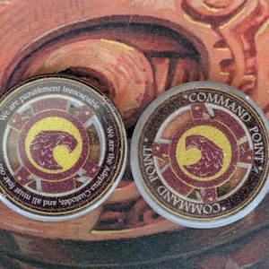 Puede incluir: Dos fichas circulares con un emblema de águila dorada y burdeos. Una dice "Command Point" y la otra tiene texto alrededor del borde que dice "We are punishment inescapable". Las fichas tienen un acabado brillante.