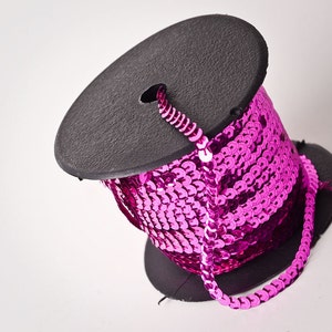Puede incluir: Un carrete de ribete de lentejuelas rosa. El ribete está enrollado alrededor de un carrete de cartón negro.