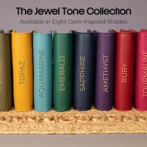 May include: The Jewel Tone Collection, featuring eight gem-inspired shades. The image displays a row of leather-bound journals in colors like Peridot, Topaz, Aquamarine, Emerald, Sapphire, Amethyst, Ruby, and Tourmaline. The journals are arranged on a woven surface.