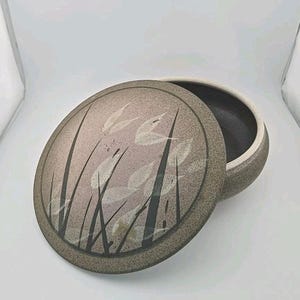May include: A round ceramic lidded container with a speckled beige and brown finish. The lid features a dark brown and cream botanical design of tall grass blades and leaves. The interior of the bowl is dark, and the rim is a light cream color.