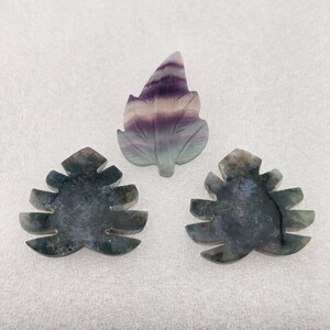 May include: Three carved stone leaf shapes. The top leaf is translucent with bands of purple, white, and green. The two bottom leaves are opaque, dark green with mottled black inclusions, featuring distinct serrated edges and pointed tips.