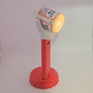 May include: A novelty flashlight featuring a red base shaped like a bottle cap, a red cylindrical handle, and a white hand-shaped mount holding a Bud Light beer can-shaped light head that emits a warm glow from the front opening.