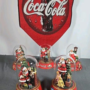 May include: A collection of Coca-Cola Christmas snow globes featuring Santa Claus figurines. Each globe sits on a red base with the Coca-Cola logo. A red Coca-Cola banner hangs in the background.
