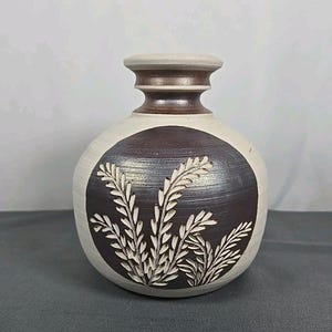 May include: A round ceramic vase with a cream-colored body and a dark brown neck. The front features a large, circular dark brown medallion with an incised, light-colored botanical pattern of wheat-like stalks carved into the surface.
