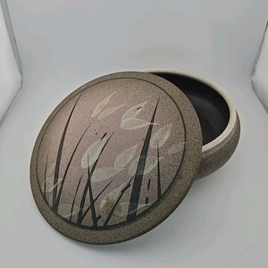 May include: A round ceramic lidded container with a speckled beige and brown finish. The lid features a decorative graphic of tall, dark grass blades and light-colored oval leaves. The interior of the container is dark, and the rim is white.