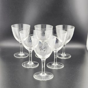 May include: Set of six clear glass wine glasses. Each glass features a delicate etched floral design on the bowl. The glasses have a classic stemmed design, perfect for serving wine or other beverages. The glasses are arranged on a dark surface.