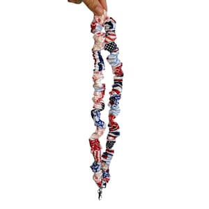 May include: A fabric lanyard featuring a gathered, scrunchie-style design with a repeating pattern of red, white, and blue stars, stripes, and paisley motifs. The long, cylindrical strap includes a metal swivel clasp at the bottom end.