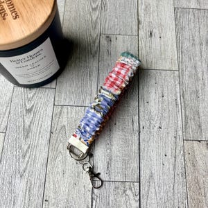 May include: A fabric wrist lanyard with a metal clasp and key rings. The lanyard is made of colorful fabric scraps in red gingham, blue floral, and other patterns. A dark blue candle with a wooden lid is in the background.
