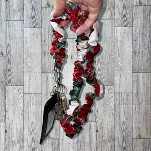 May include: A red, green, and white plaid fabric keychain with a silver metal clasp and key rings. The keychain is attached to a black wallet and several keys. The fabric is gathered and has a scrunchie-like appearance.