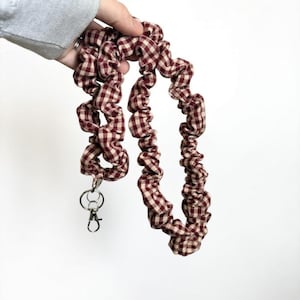 May include: A red and cream gingham pattern lanyard with a silver-colored clasp. The lanyard is made of a gathered fabric, creating a textured, ruffled appearance. The fabric is held by a hand against a white background.