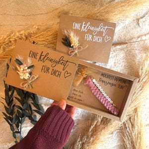Könnte beinhalten: Ein Geschenkset mit einer Kraftpapierbox mit einem rosa Makramee-Schlüsselanhänger und einer Karte. Der Schlüsselanhänger hat weiße Buchstabenperlen. Auf der Karte steht "Eine Kleinigkeit für dich" in weißer Schrift, mit einem kleinen Blumenarrangement.