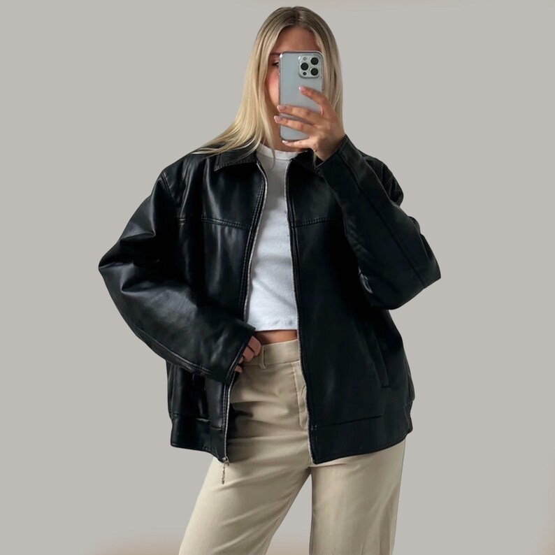 Op de afbeelding: Zwarte leren jas met kraag en ritssluiting. De jas heeft een klassiek design en een iets oversized pasvorm. Het model draagt een witte top en een beige broek.