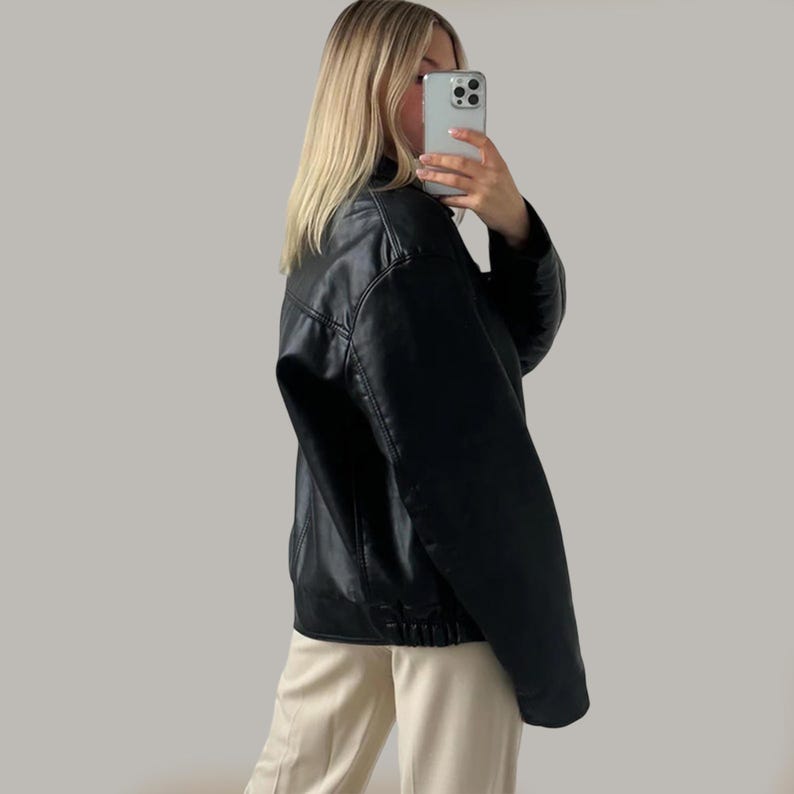 Op de afbeelding: Zwarte leren jas in bomberstijl. De jas heeft een klassieke kraag, ritssluiting en geribde manchetten en zoom. Het model draagt de jas met een beige broek.