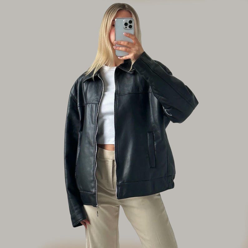 Op de afbeelding: Zwarte imitatie leren jas met een kraag, een ritssluiting en een enkele zak. De jas wordt gedragen over een witte top en gecombineerd met beige broeken. De jas heeft een klassiek, tijdloos ontwerp.