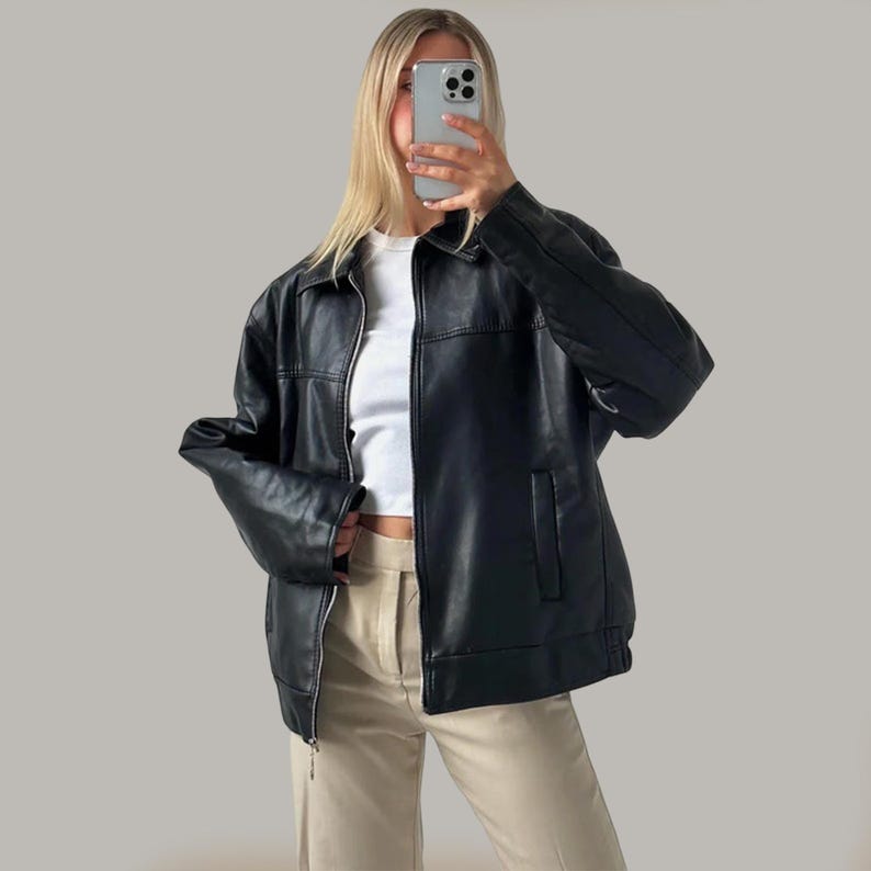 Op de afbeelding: Zwarte imitatie leren jas met een kraag, een ritssluiting aan de voorkant en een borstzak. De jas wordt gedragen over een witte top en gecombineerd met een beige broek. De jas heeft een klassieke, iets oversized pasvorm.