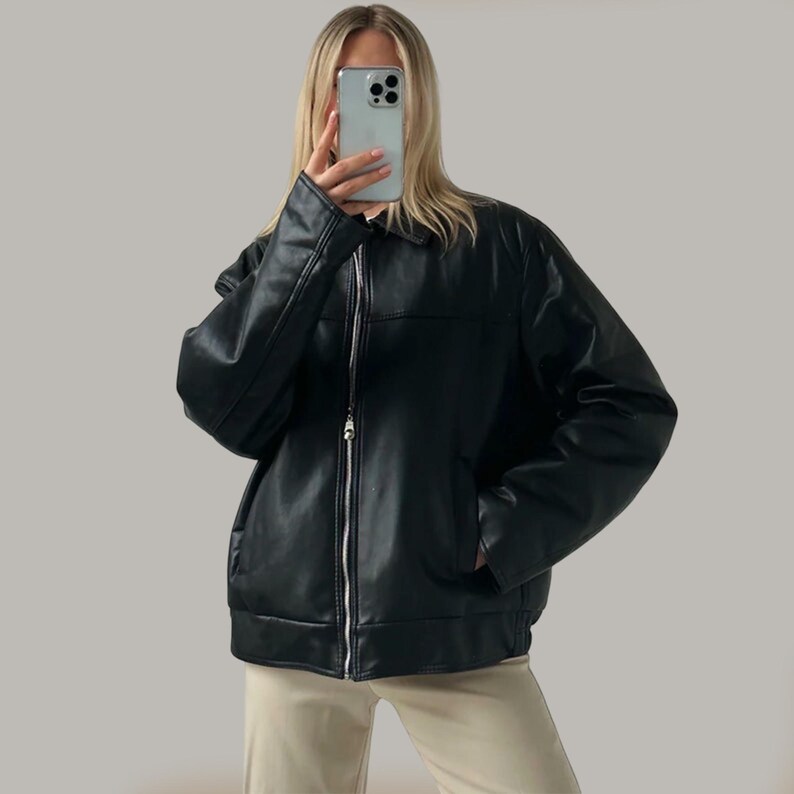 Op de afbeelding: Zwarte imitatie leren jas met een zilveren ritssluiting en twee voorzakken. De jas heeft een klassieke kraag en een iets oversized pasvorm. De jas wordt gedragen over een beige broek.