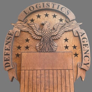 May include: A wooden plaque with the words "LOGISTICS" and "DEFENSE AGENCY" engraved on it. The plaque features a detailed carving of an eagle with outstretched wings, surrounded by stars. The plaque has a shield-shaped bottom.
