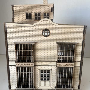 Peut inclure: Maquette en bois d'un bâtiment avec une façade en briques. Le modèle présente le texte "Ollivanders" au-dessus des fenêtres et "Makers of Fine Wands Since 382 B.C." Le bâtiment a une cheminée et une petite porte.