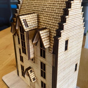 May include: A wooden model of a house with a brown roof and white walls. The house has multiple windows and a chimney. The model is sitting on a wooden base.