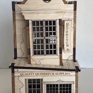May include: A detailed wooden model of a shop, featuring the text "QUALITY QUIDDITCH SUPPLIES" and a banner reading "QUIDDITCH". The building has multiple windows, a chimney, and a detailed facade. The model is made of light-coloured wood.