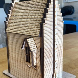 May include: A wooden model of a house with a tall chimney and a small window. The house is made of light brown wood and has a dark brown roof. The chimney is made of white bricks and has a black cap. The window is rectangular and has a white frame.