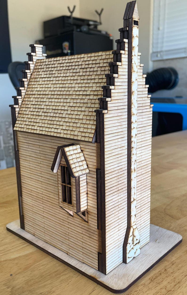 May include: A wooden model of a house with a tall chimney and a small window. The house is made of light brown wood and has a dark brown roof. The chimney is made of white bricks and has a black cap. The window is rectangular and has a white frame.