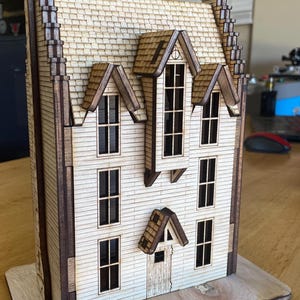 Puede incluir: Un modelo de madera de una casa con un techo marrón, revestimiento blanco y varias ventanas. La casa tiene un pequeño porche con una puerta y una ventana. El modelo está sentado sobre una base de madera.