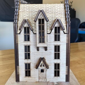 May include: A wooden model of a house with a white facade, brown trim, and a gabled roof. The house has multiple windows and a door. The model is sitting on a wooden base.
