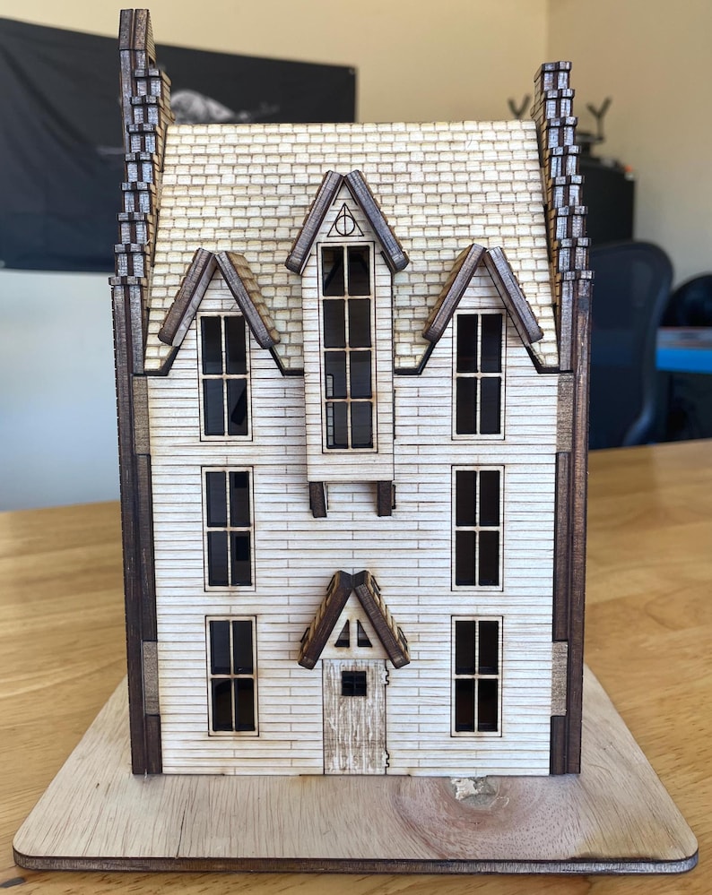May include: A wooden model of a house with a white facade, brown trim, and a gabled roof. The house has multiple windows and a door. The model is sitting on a wooden base.