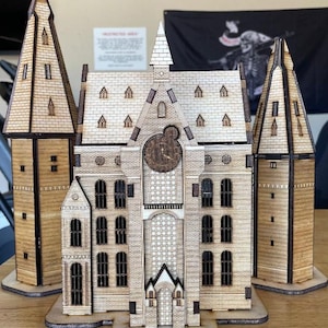 May include: A wooden model of a building with multiple towers and a clock tower. The model is made of light brown wood and features detailed architectural elements, including windows, doors, and a clock face. The model is a decorative item.
