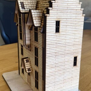 May include: A wooden model of a house with a gabled roof and multiple windows. The house is made of light brown wood and has a dark brown trim. The house is sitting on a wooden base.
