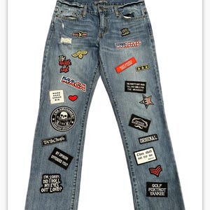 May include: Blue denim jeans with various patches, including political slogans, American flags, and other designs. The patches include "Trump", "MAGA", "The Hustle Is Real", "Work Hard Dream Big", "2nd Amendment", "We The People", "My Opinion Offended You", "I'm Sorry Did I Roll My Eyes Out Loud", "You're Perfect", "Make America Great Again", "The Party's Not Over Till You Smile For The Mugshot", "Try Your Kindness Challenge", "Original", "Chou Shall Not Try Me", "Golf Foxtrot Yankee".