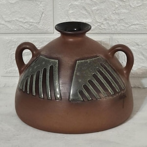 May include: Brown ceramic vase with two handles and a dark rim. The vase features a symmetrical design of green and gray geometric shapes on the front. The vase has a rounded body and a narrow neck.