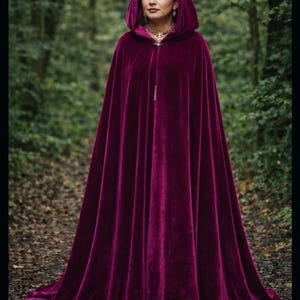 May include: A long, hooded velvet cloak in a deep burgundy color. The cloak drapes to the ground and features a gold clasp at the neckline. The person wearing the cloak is standing in a forest setting.
